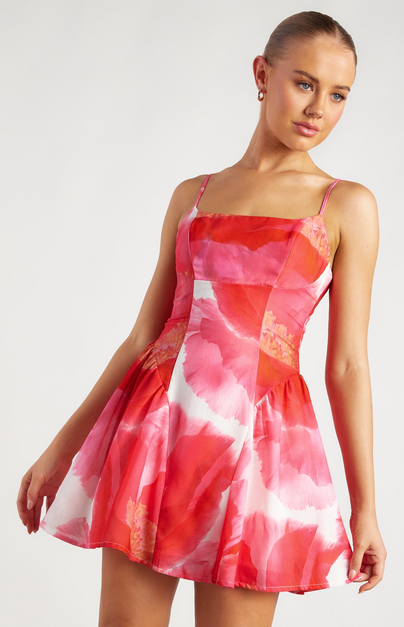 The coco dress is a vibrant and feminine mini dress featuring a striking pink and red floral pattern. It has a fitted bodice with a structured silhouette, accentuating the waist before flaring into a short, flouncy A-line skirt. The bodice is designed with delicate spaghetti straps, adding a touch of elegance and lightness to the overall look.
The neckline is slightly asymmetrical, with an angular panel that runs diagonally across the front, creating a unique and modern design element. The fabric a