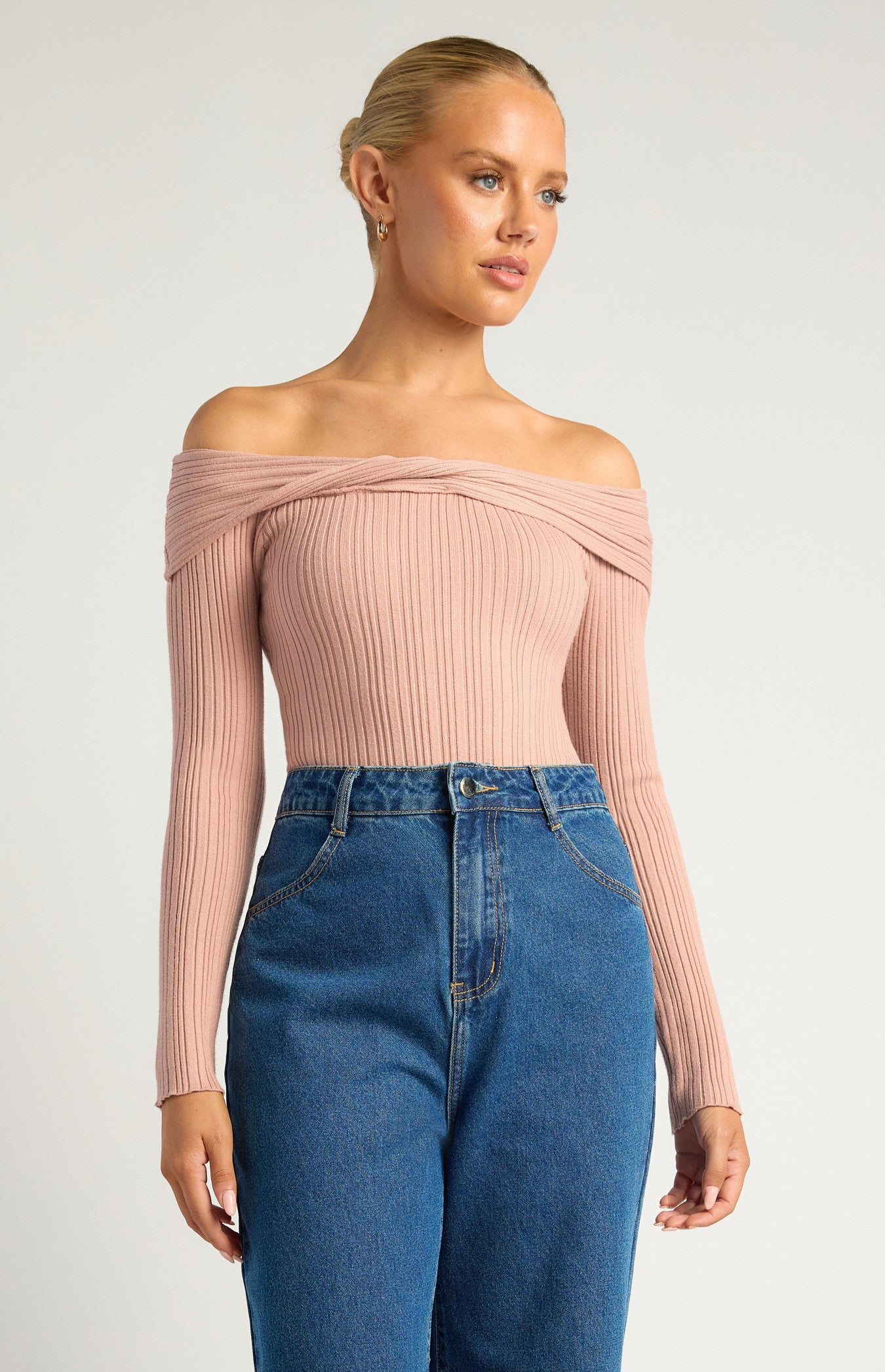 This is a long-sleeve, off-the-shoulder bodysuit designed to hug the body comfortably. The neckline is a twist-front, off-the-shoulder style, giving it a feminine, elegant look while showcasing the collarbones. The colour is a soft, muted blush pink — neutral enough to pair easily with other wardrobe pieces