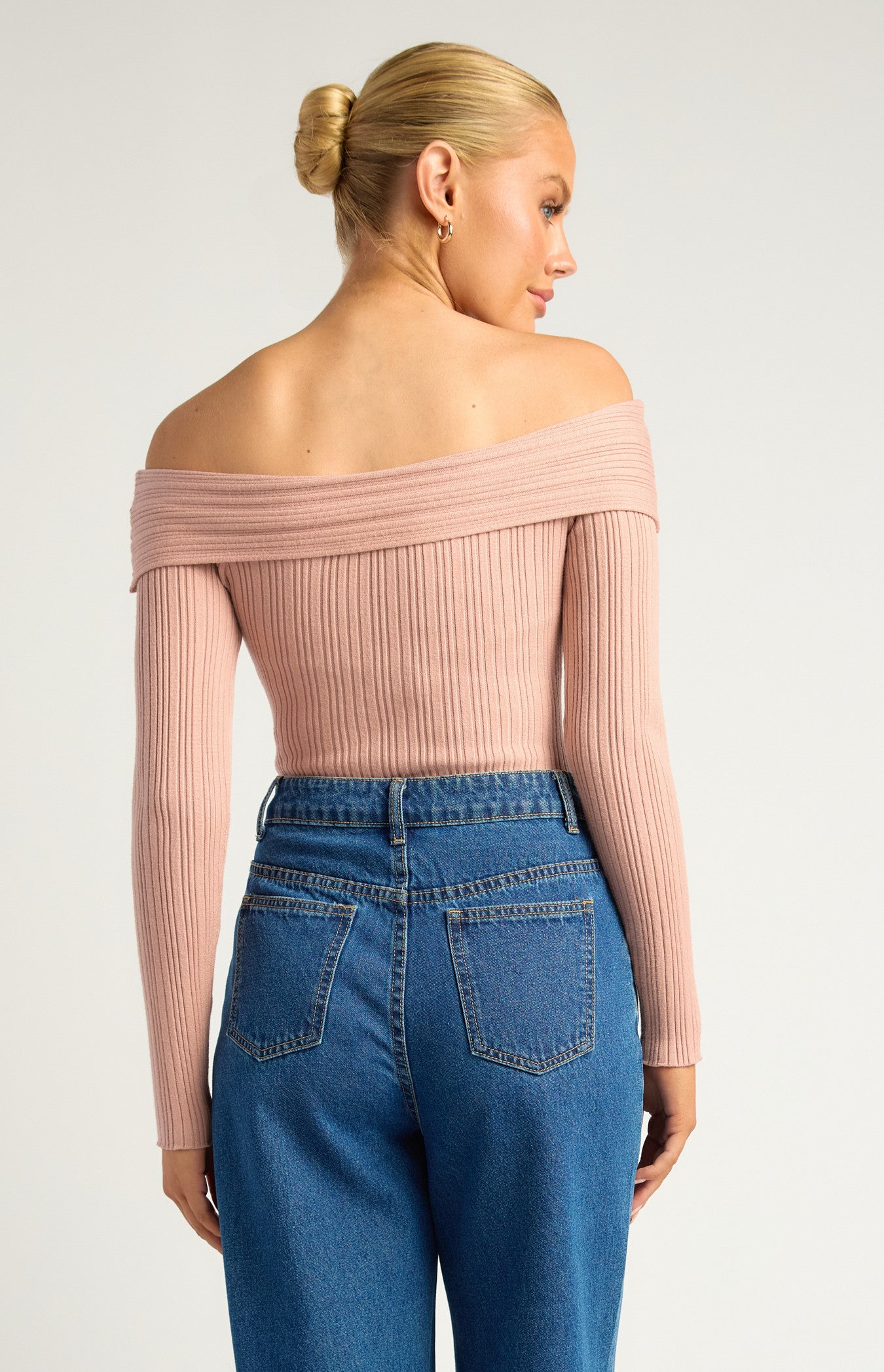 This is a long-sleeve, off-the-shoulder bodysuit designed to hug the body comfortably. The neckline is a twist-front, off-the-shoulder style, giving it a feminine, elegant look while showcasing the collarbones. The colour is a soft, muted blush pink — neutral enough to pair easily with other wardrobe pieces