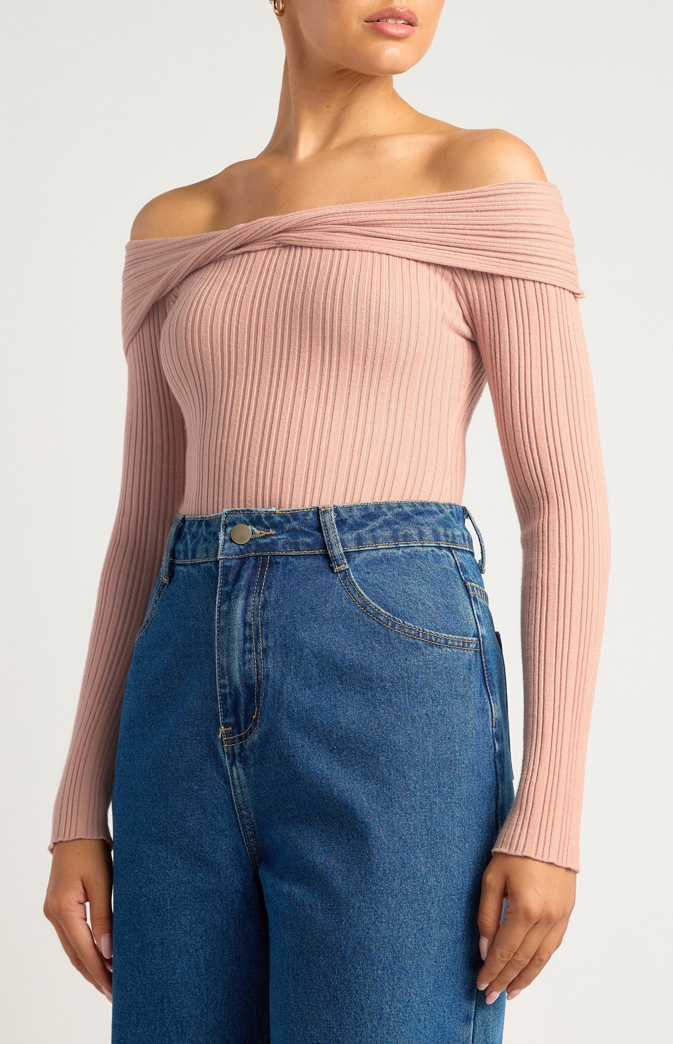 This is a long-sleeve, off-the-shoulder bodysuit designed to hug the body comfortably. The neckline is a twist-front, off-the-shoulder style, giving it a feminine, elegant look while showcasing the collarbones. The colour is a soft, muted blush pink — neutral enough to pair easily with other wardrobe pieces