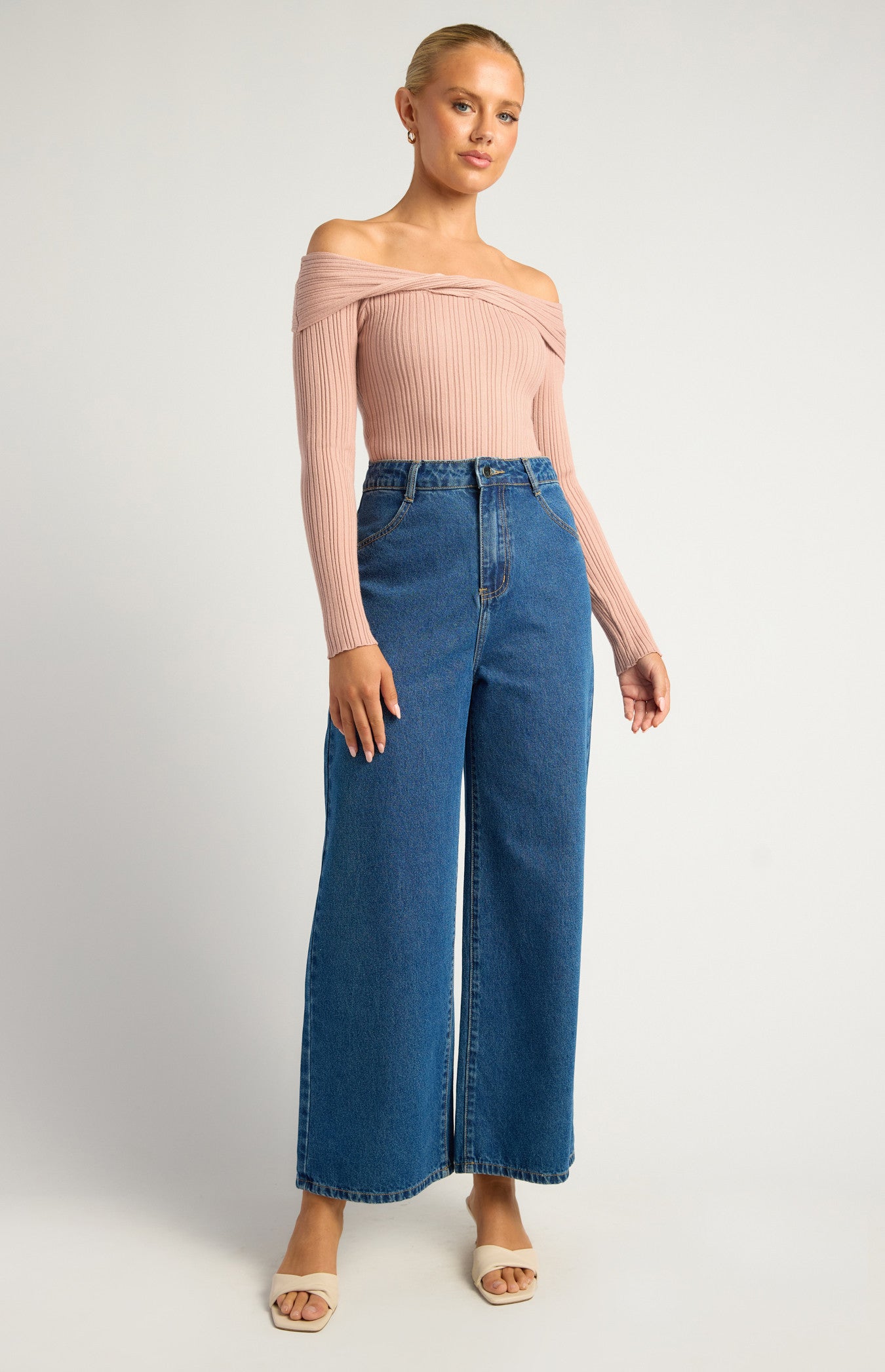 This is a long-sleeve, off-the-shoulder bodysuit designed to hug the body comfortably. The neckline is a twist-front, off-the-shoulder style, giving it a feminine, elegant look while showcasing the collarbones. The colour is a soft, muted blush pink — neutral enough to pair easily with other wardrobe pieces