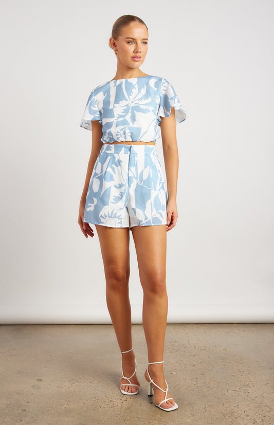 Esther Two-Piece Shirt and Crop Set
	•	Top: Reversible design with a crossover section at the front or back, adding flexibility to your look. It features an elasticized waistband at the bottom, cutting the top to mid-length with short sleeves. The fabric is a pale blue with a geometric tropical pattern of palm trees, leaves, and suns, giving it a fresh, sunny vibe.
	•	Shorts: High-waisted, zip-up d