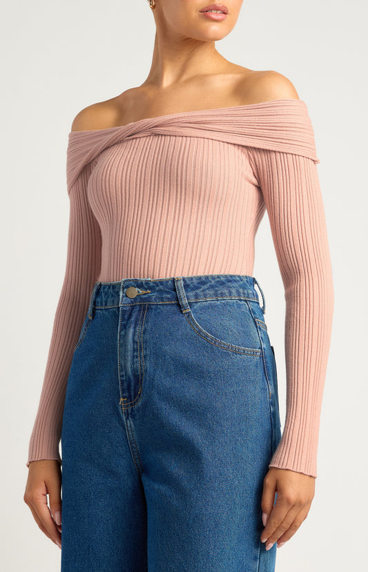 This is a long-sleeve, off-the-shoulder bodysuit designed to hug the body comfortably. The neckline is a twist-front, off-the-shoulder style, giving it a feminine, elegant look while showcasing the collarbones. The colour is a soft, muted blush pink — neutral enough to pair easily with other wardrobe pieces